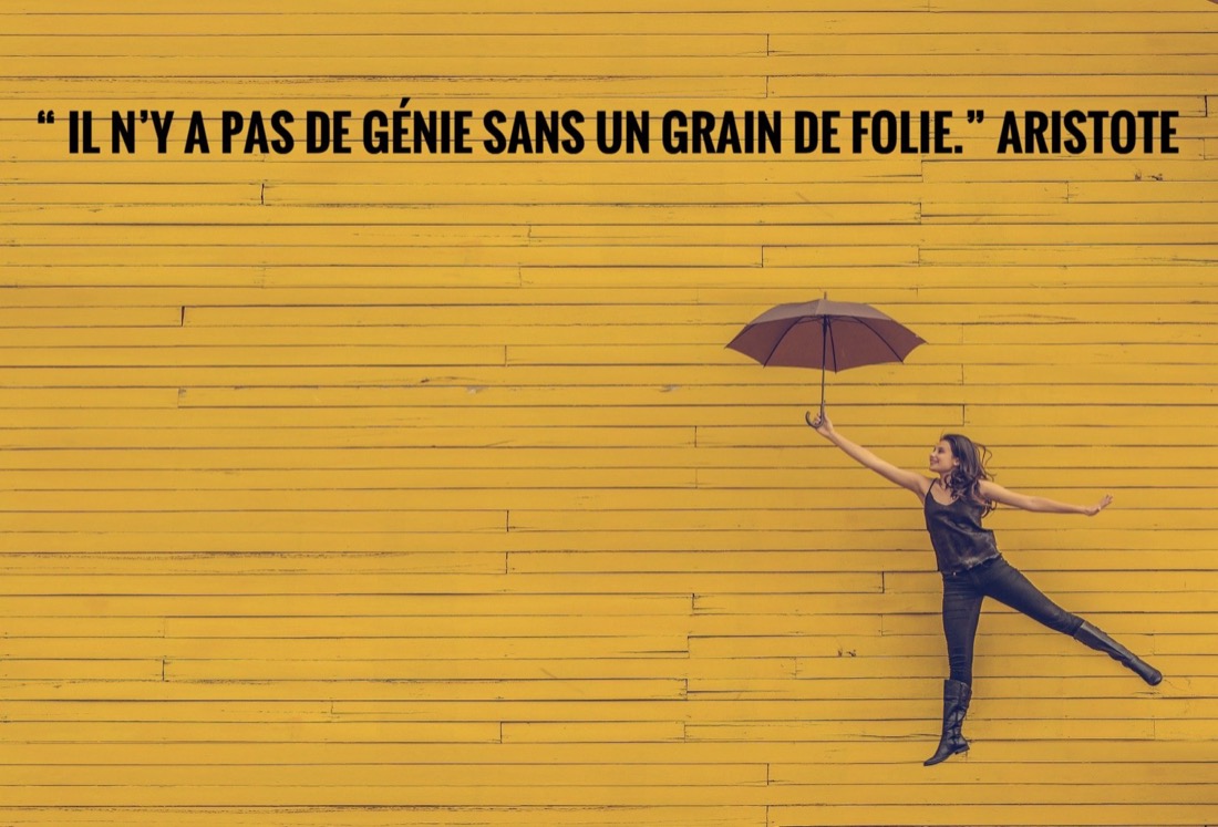 Femme bondissant avec un parapluie devant un mur jaune, citation d'Aristote sur le grain de folie.